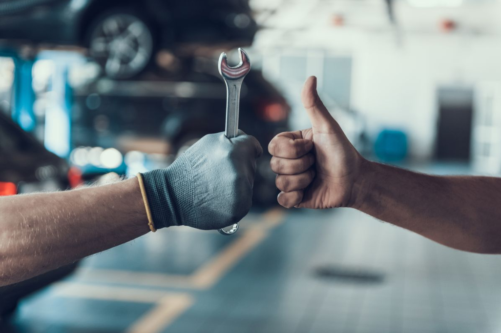 two hands fist bumping, one is gloved and holding a wrench and one is giving a thumbs up sign.