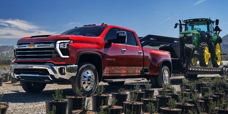 Red Chevrolet Silverado HD pickup truck towing a trailer with agricultural equipment, parked outdoors with mountains in the background, highlighting heavy-duty towing capability.